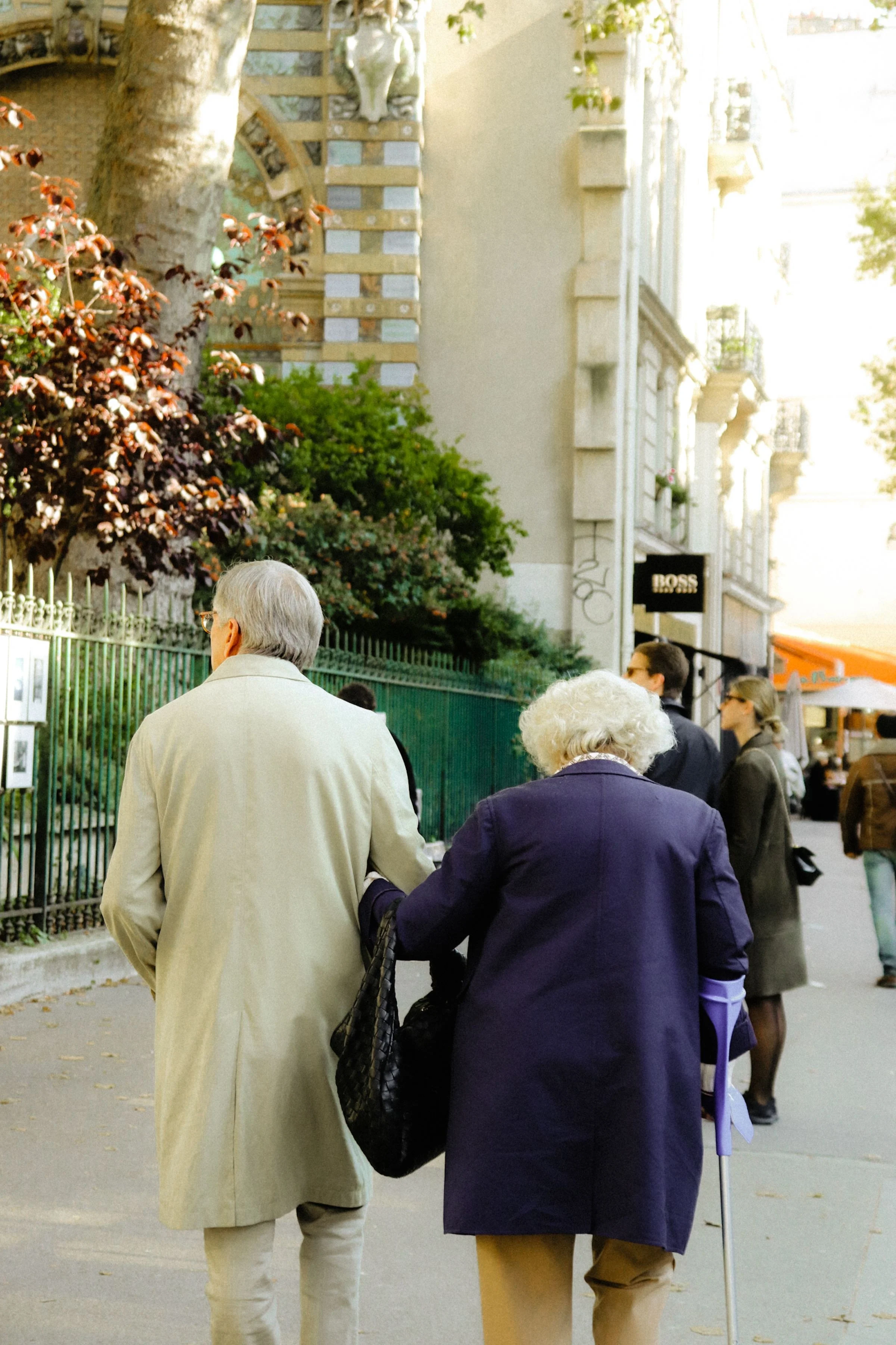 Casal de idosos caminhando de braços dados numa calçada arborizada, com bengala apoiada do lado saudável