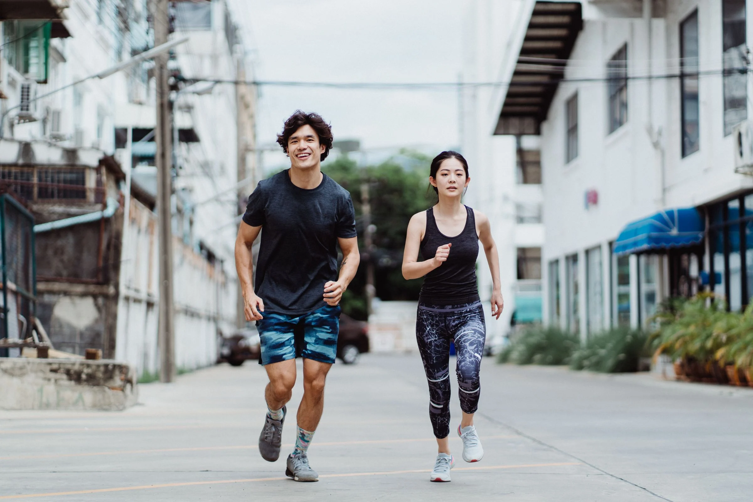 Casal correndo lado a lado numa rua, de corpo inteiro, em roupa esportiva e tênis de corrida