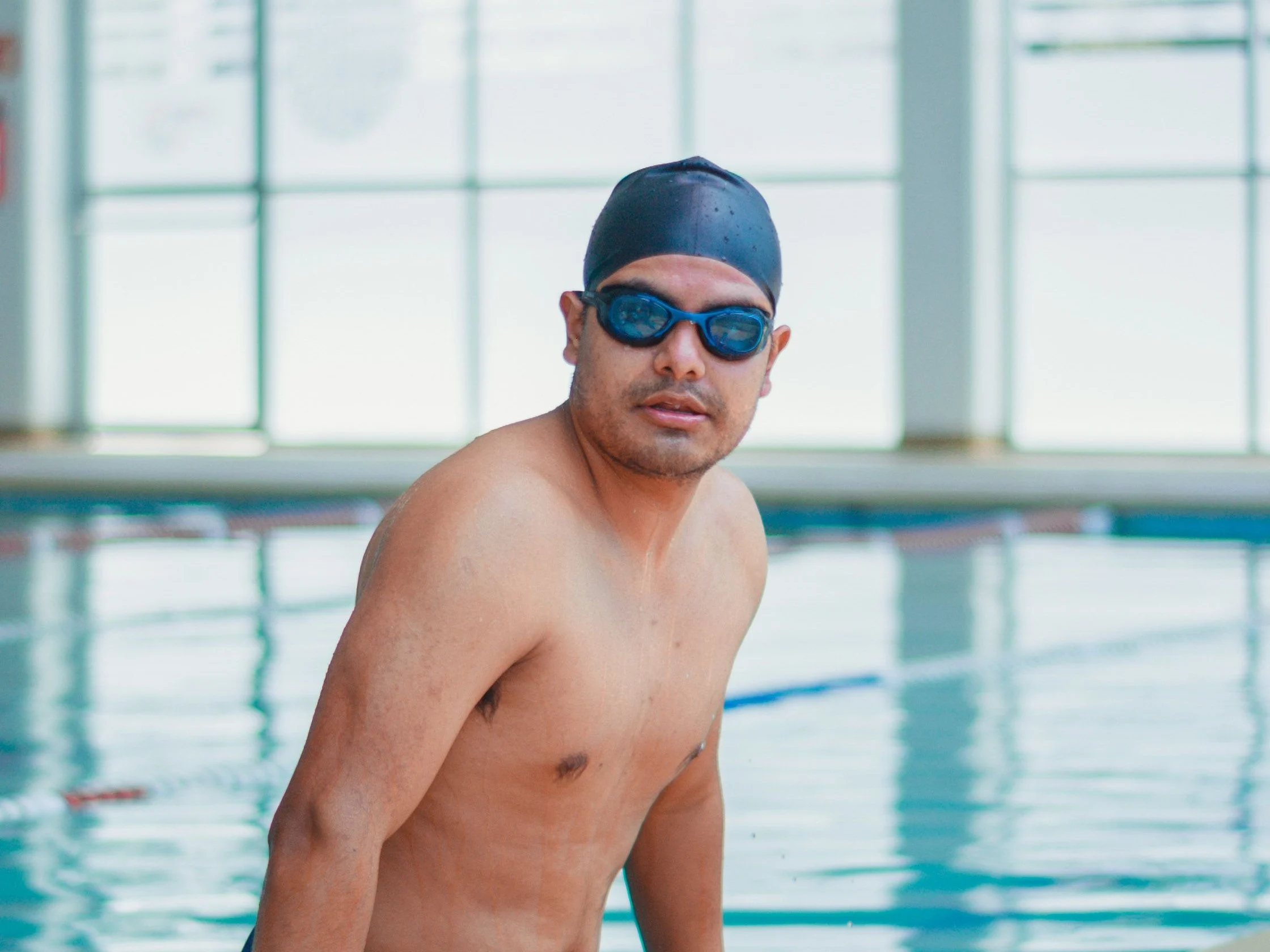 Homem à beira de uma piscina coberta, de touca e óculos de natação, apoiado na borda