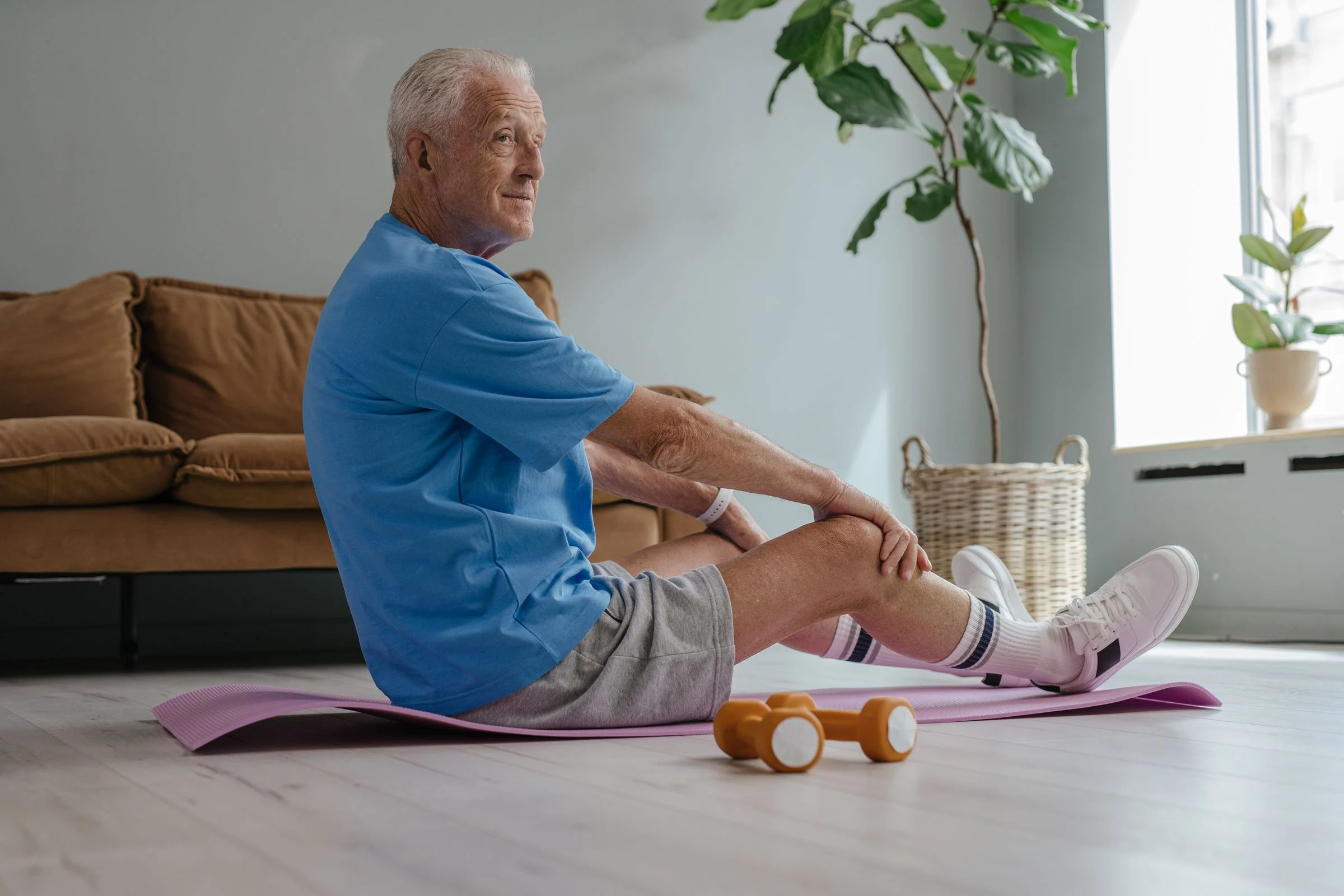 Homem idoso sentado num tapete de exercício em casa, alongando a perna, com halteres leves ao lado