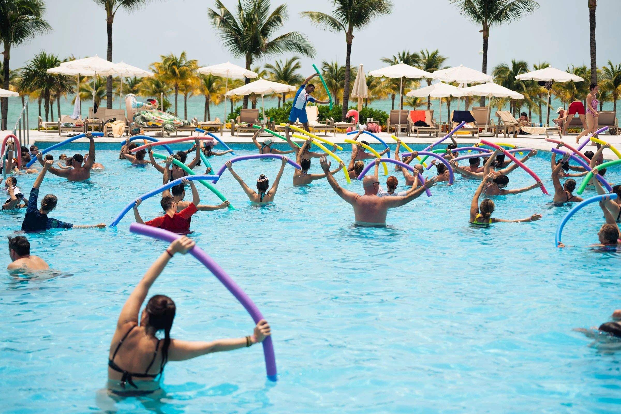 Turma de hidroginástica numa piscina ao ar livre, com os participantes erguendo espaguetes de espuma acompanhando o instrutor