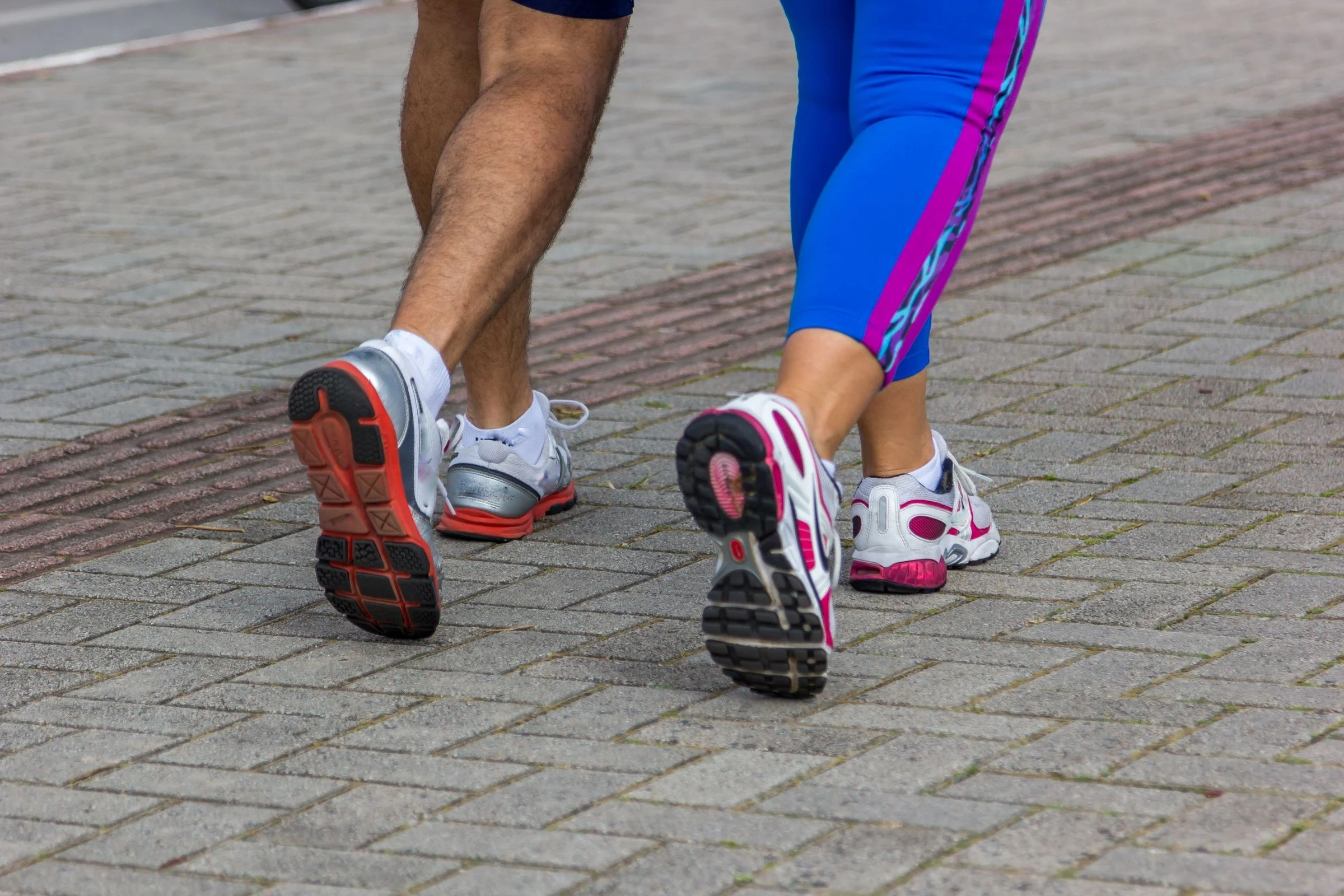 Pernas e pés de duas pessoas caminhando lado a lado, em tênis esportivos, sobre calçada de pedra